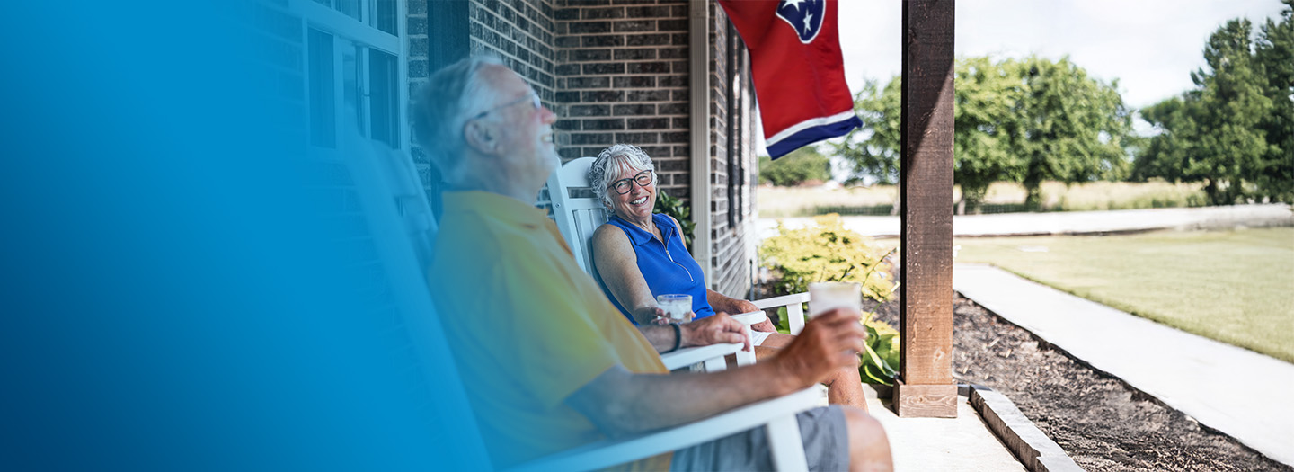 An older couple conversing and smiling while sitting on their front porch in rocking chairs.